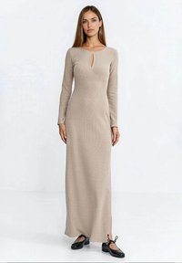 Beżowa, długorękawowa, prążkowana sukienka z wycięciem w kształcie łezki przy dekolcie i bocznymi rozcięciami, zestawiona z czarnymi płaskimi butami.
