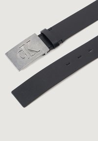 Curea din piele neagră cu o cataramă rectangulară argintie care are gravat logo-ul "CK" și o bandă netedă, plată, cu găuri pentru ajustare.