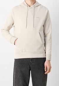 Felpa beige chiara con tasca frontale, cappuccio con cordino e un delicato dettaglio del logo; realizzata in tessuto morbido e liscio; abbinata a jeans scuri.