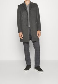 Abrigo de lana gris con tres botones, solapas de muesca y dos bolsillos frontales, combinado con pantalones grises texturizados y zapatillas negras.