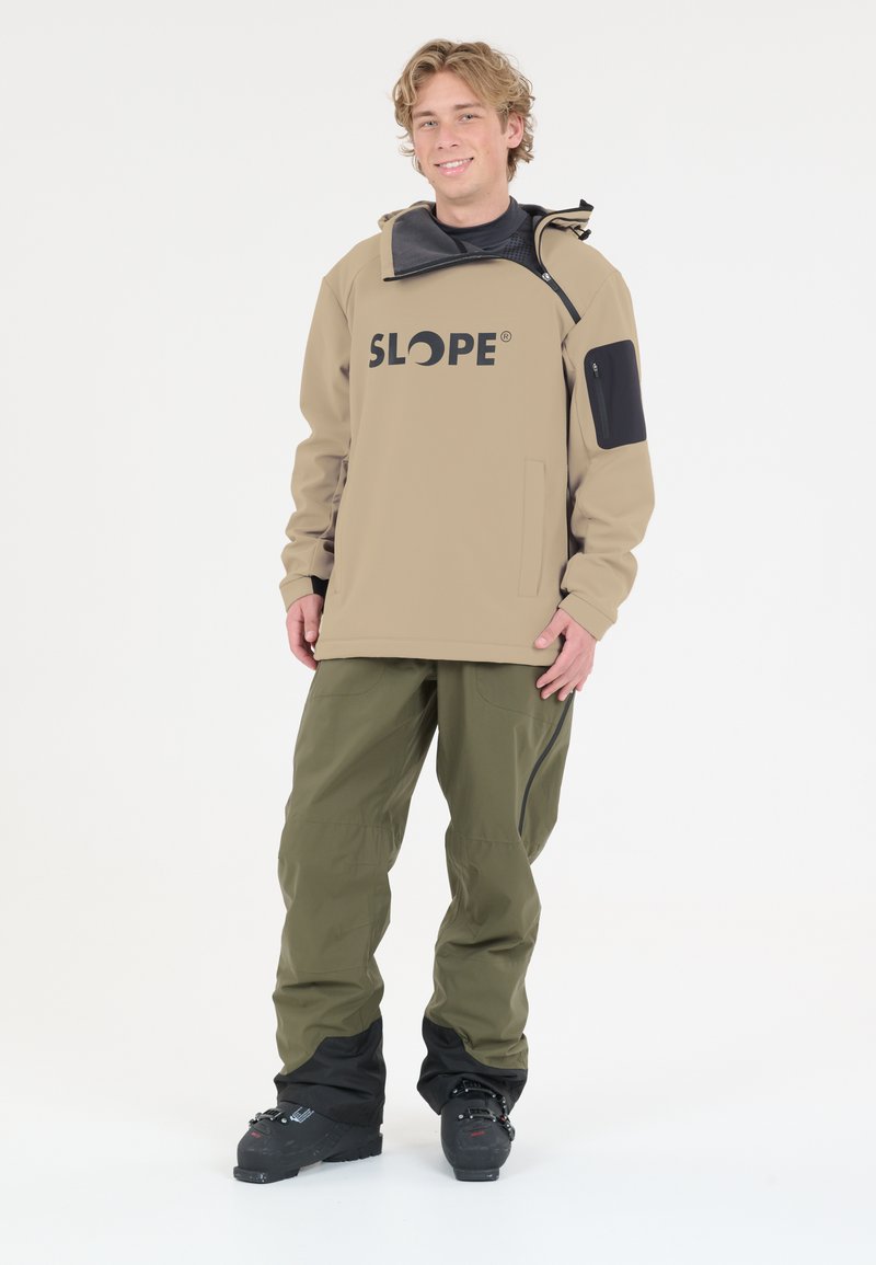 Veste imperméable beige avec un logo noir, dotée d'une poche zippée et d'accents noirs contrastants, associée à un pantalon vert olive et des bottes noires.