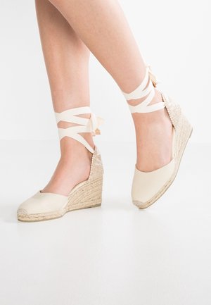 Keil-Espadrilles mit cremefarbenem Stoff, geflochtenem Jutesohlenabsatz und an den Füßen einer Person mit Schleifen gebundenen Knöchelriemen.