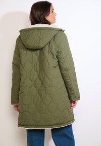 Žena v olivovo zelenej prešívanej kapucňovej bunde s bielou flísovou podšívkou, zobrazená zozadu, v kombinácii s modrými džínsami.