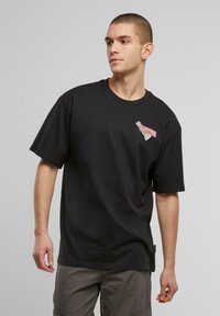 Black cotton t-shirt featuring a colorful graphic patch on the left chest. Short sleeves, crew neck, relaxed fit.