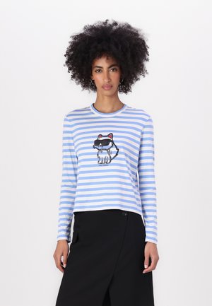 Chemise à manches longues rayée bleu clair et blanche, avec un motif de chat dans des tons de gris, noir et rouge au centre, et un col rond.