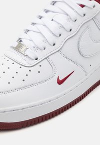 Biele kožené tenisky s perforovanou špičkou, červeným logom Nike, striebornou hardvérovou nášivkou a textúrovanou červenou gumovou podrážkou. Šnúrky sú biele a ploché.