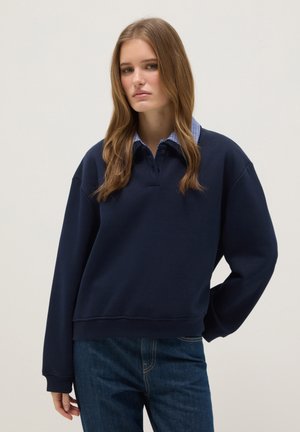 Jonge vrouw met lang haar die een marineblauwe trui draagt over een blauw geruit overhemd met kraag en blauwe spijkerbroek, staand voor een effen achtergrond.