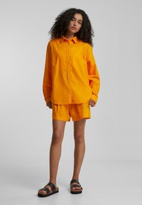 Bright orange linen shirt and shorts with a relaxed fit. Paired with black platform sandals featuring two wide straps. Smooth texture.