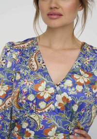 Bloemen wikkeljurk in blauwe stof met levendige oranje, groene en witte patronen. Heeft een V-halslijn en korte pofmouwen. De textuur is glad.