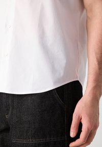 Balta marškiniai su mygtukais, minkšta tekstūra, trumpomis rankovėmis ir apvaliu priekiu, derinami su tamsių džinsų kelnėmis, turinčiomis kontrastingą siuvinėjimą.