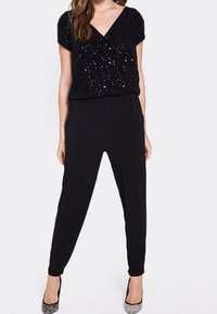 Mono negro con cintura ajustada, mangas cortas y escote en V; parte superior adornada con lentejuelas, piernas ajustadas y tacones metálicos.