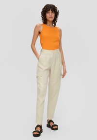 Oranje mouwloze tanktop gecombineerd met beige cargobroek met zijzakken en zwarte chunky sandalen op een neutrale achtergrond.
