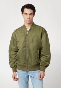 Jonge man met donker krullend haar, gekleed in een olijfgroene bomberjack met subtiele bloemdessin en lichtblauwe jeans, staande tegen een witte achtergrond.