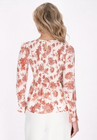 Blusa floreale con maniche lunghe, caratterizzata da una apertura a chiave sul retro, texture arricciata, di colore crema con motivi floreali rossi. Tessuto morbido e leggero.