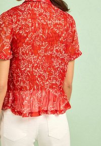 Blouse rouge transparente avec un imprimé floral, manches courtes et un ourlet peplum froncé, stylée avec un pantalon blanc.
