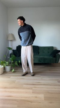 Grau und schwarz gefütterter Reißverschluss-Pullover mit hohem Kragen, kombiniert mit lockeren hellen Hosen und beigen Schuhen, vor einer grünen Couch und Zimmerpflanzen.