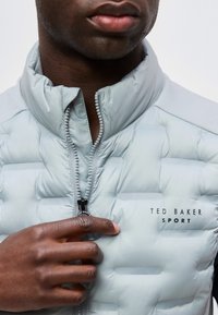 Κοντινό ενός ατόμου που φοράει ανοιχτογκρι παπλωμένη ζακέτα Ted Baker Sport, μερικώς φερμουαρισμένη, χέρι αγγίζει την περιοχή του φερμουάρ.