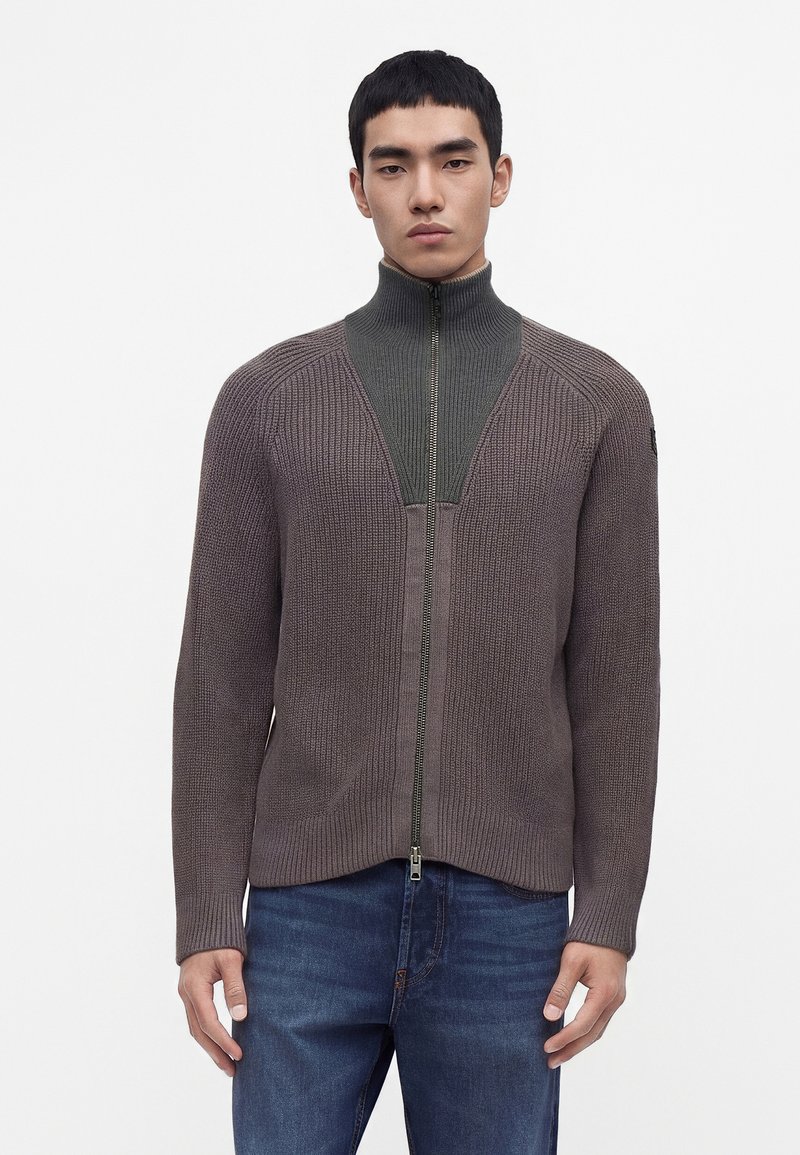 Jeune homme portant un pull marron tricoté zippé avec un col montant et un jean bleu, debout devant un fond blanc uni.