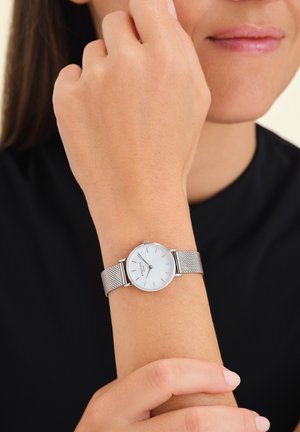Montre-bracelet en argent avec un cadran blanc, design minimaliste, bracelet en maille et marqueurs d'heure discrets, portée sur une main contre un fond sombre.