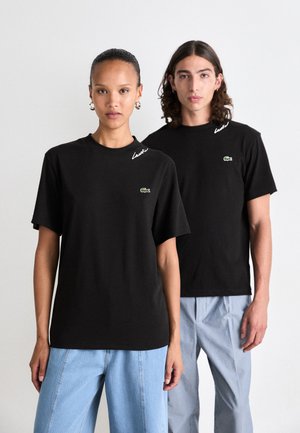 Black cotton t-shirts with crew necks, featuring embroidered white text and a small green logo on the chest. Worn with loose blue denim and striped pants.