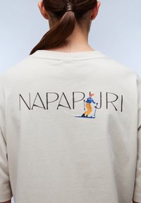 Beige bomulds t-shirt med et trykt grafisk motiv af en skihopper i blå og gul, med "NAPAPIJRI" i fede sorte bogstaver ovenfor.