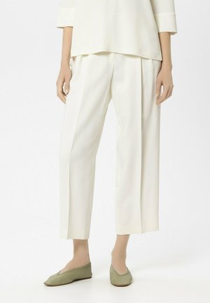 Frau trägt cremefarbene, weit geschnittene, knöchellange Hose, cremefarbenes Oberteil und olivgrüne flache Schuhe, steht vor weißem Hintergrund.