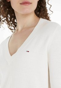 Maglione bianco a maglia con scollo a V e dettagli a coste; piccola toppa con logo rosso, bianco e blu sul lato sinistro del petto.