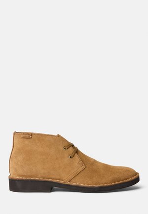 Hellbrauner Wildleder-Knöchelboot mit Zwei-Loch-Schnürsenkeln, schwarzer Sohle und kleinem, angenähtem Polo-Label nahe der Ferse.