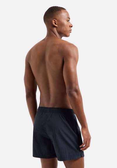 Costume da bagno nero con vita elastica, tessuto testurizzato e spacchi laterali, indossato su schiena nuda su uno sfondo chiaro.