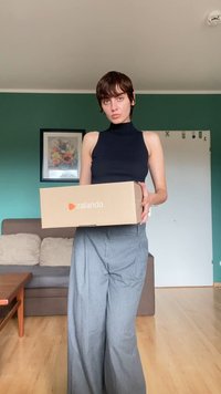 Egy Zalando kartondoboz, amelyet egy fekete ujjatlan felsőt és szürke bővebb nadrágot viselő személy tart a kezében, a háttérben egy zöld fal és kanapé látható.