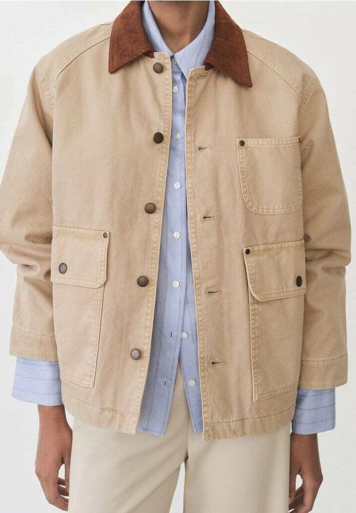 Veste beige avec col marron, fermeture à boutons à l’avant et plusieurs poches, portée sur une chemise bleu clair boutonnée et un pantalon beige.