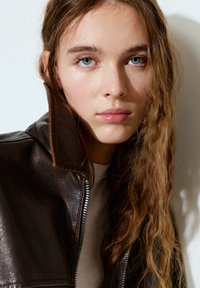 Braune Lederjacke mit hohem Kragen und sichtbarem Reißverschluss, getragen über einem hellen Hemd. Verfügt über eine weiche Textur und eine glatte Oberfläche.