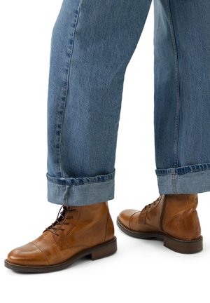 Bottines en cuir marron avec lacets, bout arrondi et finition texturée. Portées avec un jean en denim retroussé, mettant en valeur des couleurs contrastantes.