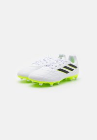 Botas de fútbol blancas con un acabado texturizado, que presentan rayas negras y suelas de un verde brillante. Diseño con cordones y clavos moldeados para tracción.