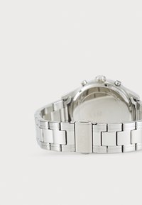 Ασημένιο ρολόι χειρός από ανοξείδωτο ατσάλι με βραχιόλι από συνδέσμους, στρογγυλή κάσα και τρία κουμπιά στην πλευρά. Η πίσω πλευρά είναι σκαλισμένη με την ένδειξη "TIMEX."