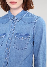 Ljusblå denimskjorta med klassisk krage, två framfickor och silverfärgade tryckknappar. Synliga sömmar och mjuk textur.