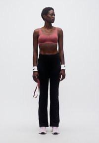 Γυναίκα με ροζ αθλητικό σουτιέν, μαύρο κολάν, λευκά περιβραχιόνια και ροζ αθλητικά παπούτσια, στέκεται με λάστιχο αντίστασης πάνω σε λευκό φόντο.