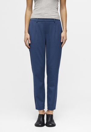 Kvinde iført blå skræddersyede ankellange bukser, grå ærmeløs top og sorte slip-on sko, stående mod en ensfarvet lys baggrund.