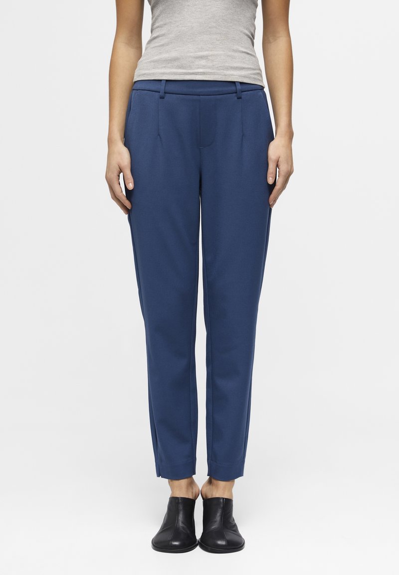 Femme portant un pantalon bleu ajusté aux chevilles, un haut gris sans manches et des chaussures slip-on noires, debout devant un fond clair uni.