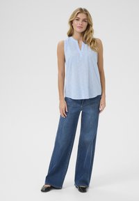 Blouse sans manches bleu clair avec un col en V, motif texturé, associée à un pantalon large en denim bleu foncé et des chaussures noires.