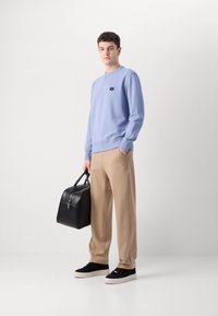 Lyseblå sweatshirt med rund hals og sort label, brune vide bukser, sorte høj-top sneakers og en sort lædertaske med sølvhardware.