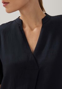 Navyblaue Bluse mit V-Ausschnitt und kurzen Ärmeln, aus glattem Stoff mit einer subtilen Textur und klaren Linien im Design.