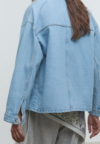 Personne portant une veste en denim bleu clair sur une chemise à motif floral et un pantalon gris, vue de dos et de la taille vers le bas.