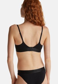 Žena s vlnitými rudými vlasy, oblečená v černé podprsence a odpovídajícím spodním prádle, zobrazená zadečky proti jednoduchému bílému pozadí.