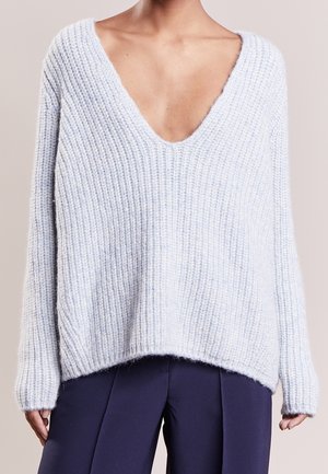 Mujer con un suéter grueso de punto color azul claro, con escote pronunciado en V y ajuste holgado, sobre pantalones azul oscuro.