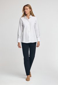 Witte langpols overhemd met knopen aan de voorkant, gecombineerd met donkere broek en beige instappers. Glad stof met klassieke kraag en omgeslagen mouwen.