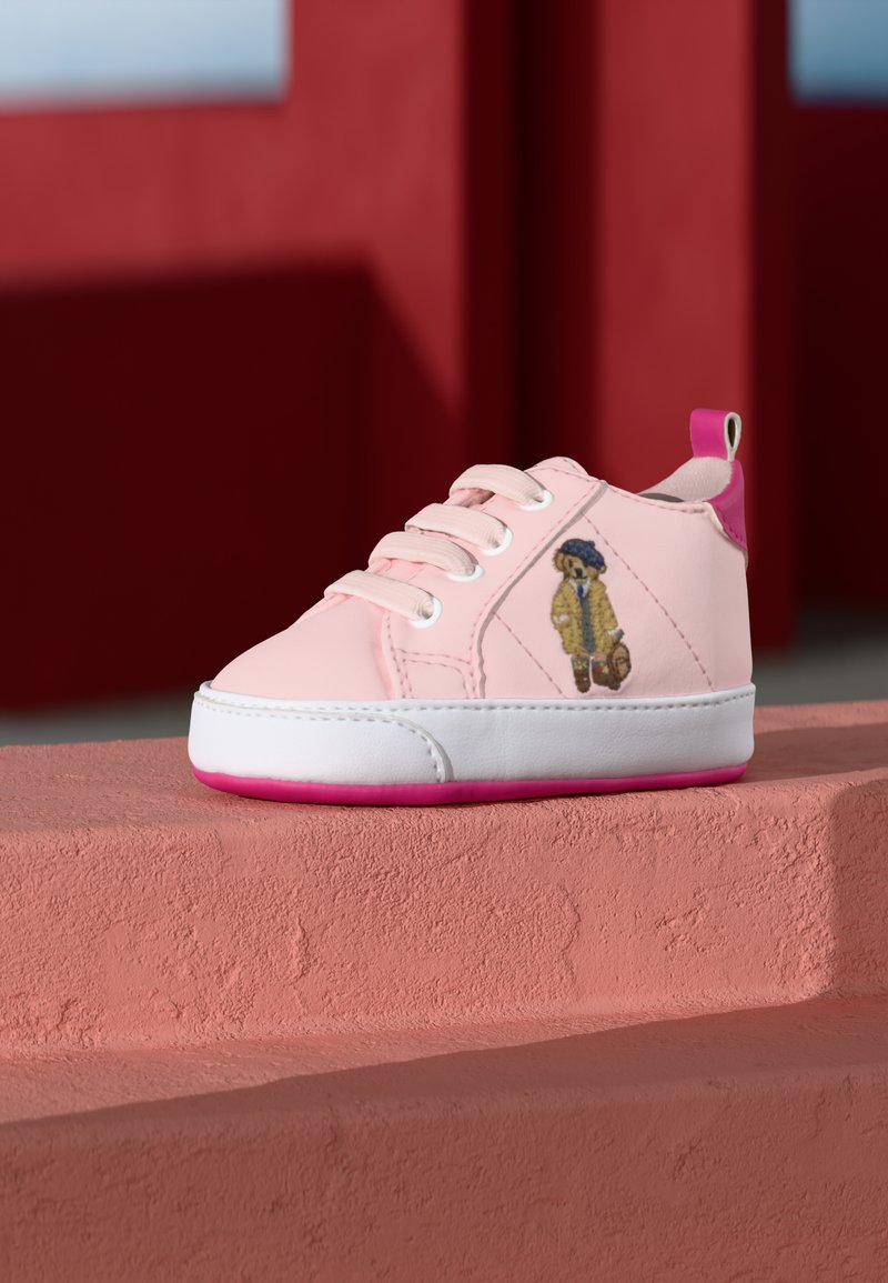 Sapatilha rosa com sola branca e detalhes em rosa. Apresenta atacadores brancos e um design de urso bordado na lateral. Textura suave.