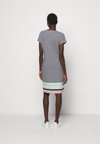 Kobieta stoi, ubrana w sukienkę z krótkim rękawem, do kolan, w czarno-białe paski oraz pastelowe poziome pasy na dole, w białych sneakersach.