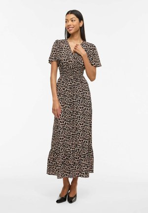 Kobieta w midi sukience w panterkę z krótkimi rękawami i czarnych szpilkach, stojąca i uśmiechająca się, z jedną ręką przy szyi.