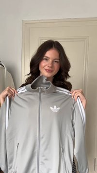 Gray zip-up jacket made of smooth fabric with white Adidas logo and three white stripes on sleeves. Features side pockets and high collar.